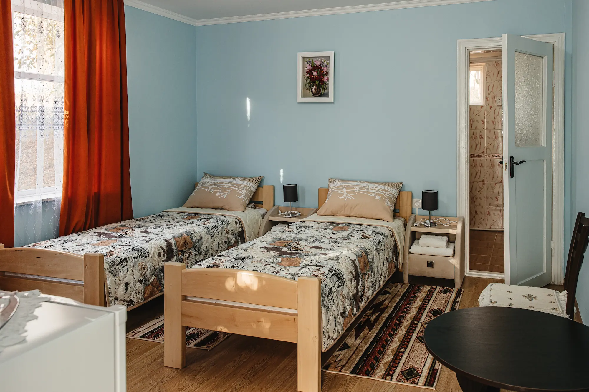 Cameră cu două paturi de o persoană din lemn deschis, pereți bleu, draperii cărămizii și ușa deschisă spre baie
