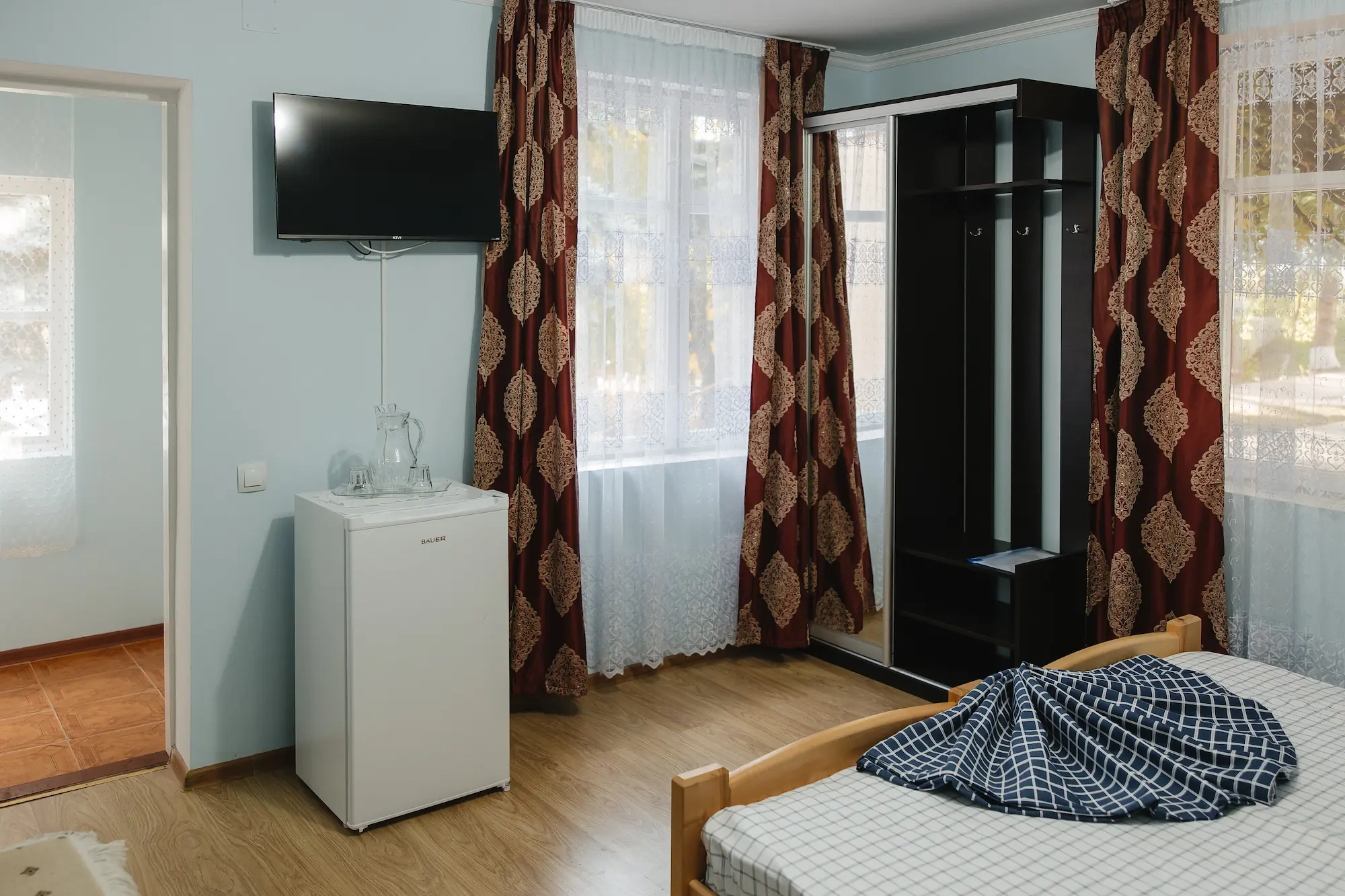 Colț de cameră cu televizor pe perete, frigider mic cu carafă și pahare deasupra, dulap și draperii cu model