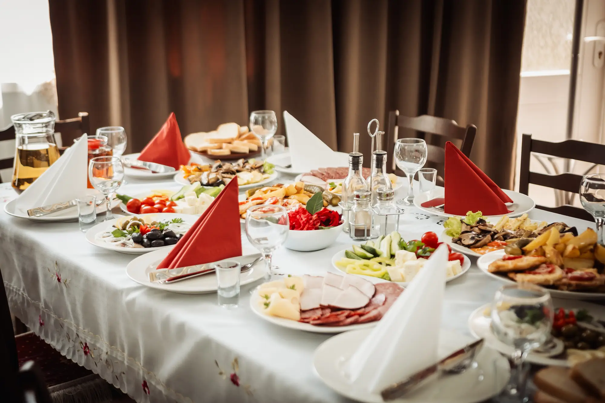 Masă întinsă cu aperitive reci, brânzeturi, legume proaspete și pâine de casă, cu șervete roșii și pahare pregătite