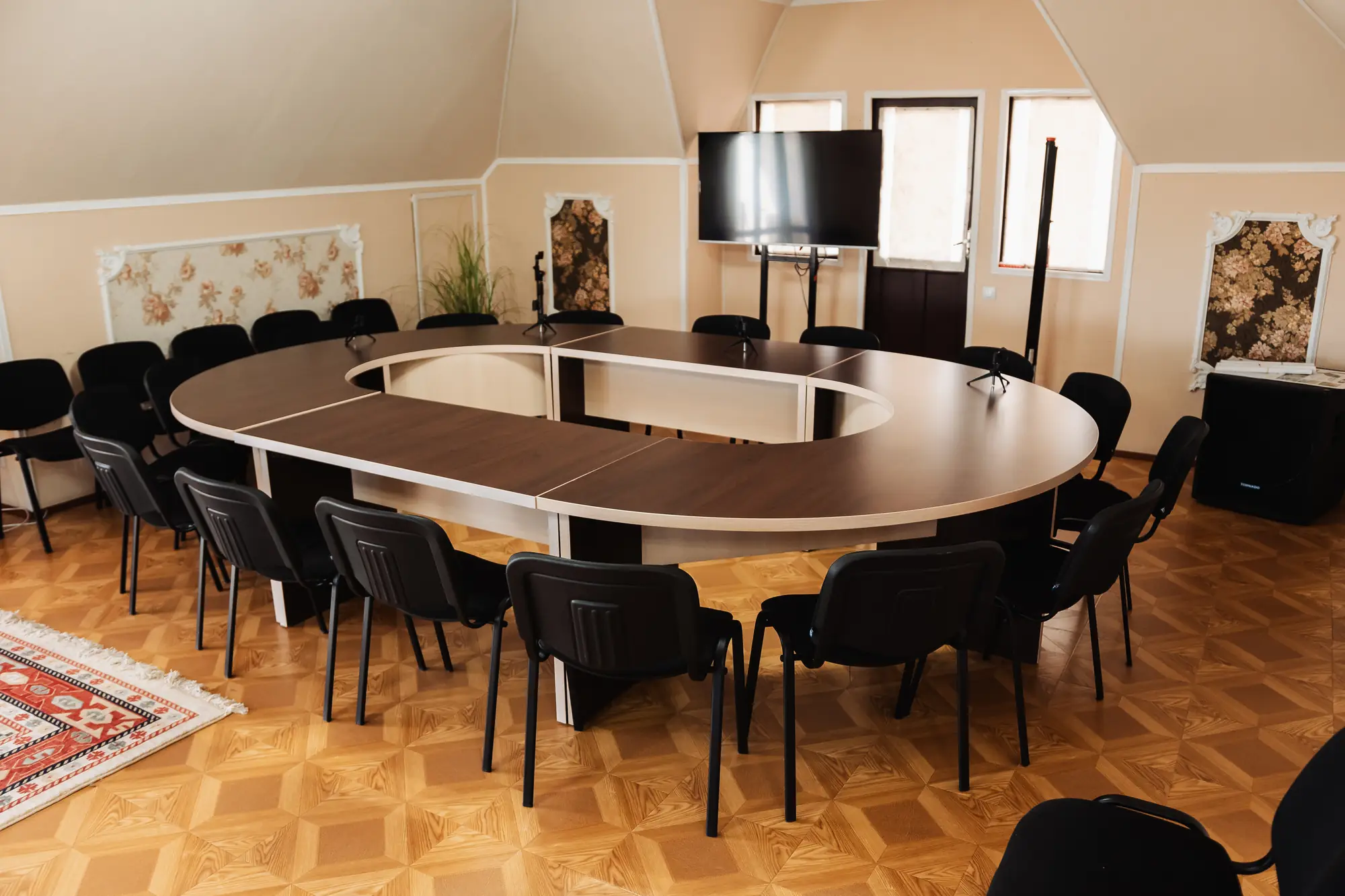 Sala de conferințe cu masă ovală, scaune de jur împrejur, microfoane și televizor mare pe suport