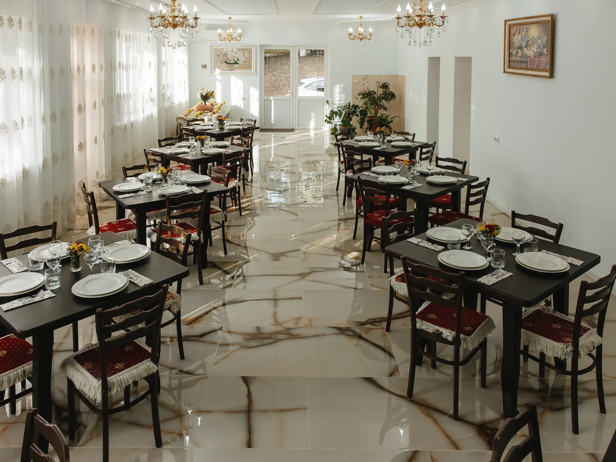 Sala restaurantului cu mese de lemn închis aranjate pentru masă, scaune cu perne roșii, candelabre de cristal și lumină naturală prin perdele